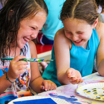 Two girls sitting at a table painting and laughing.